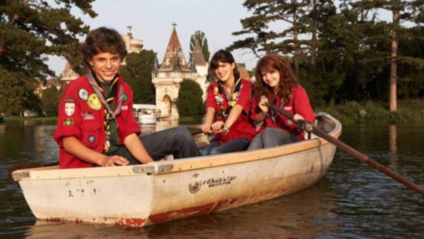 Drei Pfadfinder in roten Hemden rudern auf einem See vor einem Schloss.