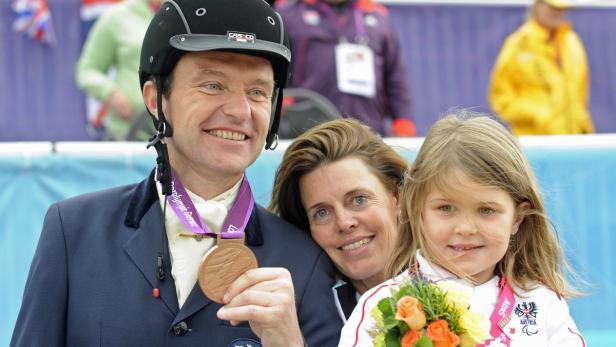 Ein Mann mit Reithelm und Bronzemedaille posiert mit einer Frau und einem kleinen Mädchen mit Blumenstrauß.