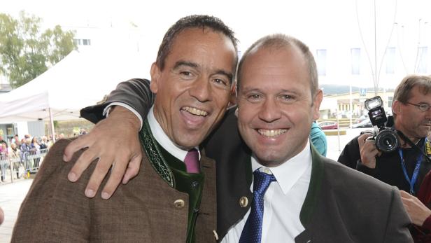 Zwei Männer in Trachtenjacken umarmen sich und lächeln in die Kamera.