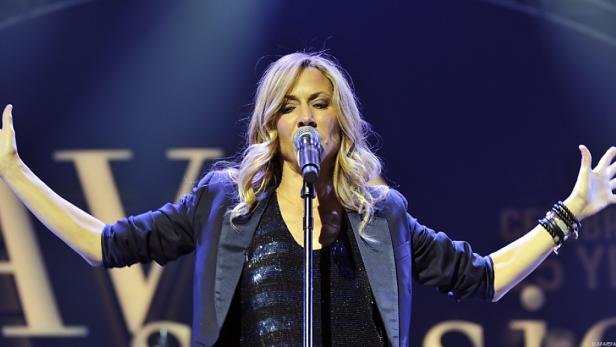 Sheryl Crow singt mit erhobenen Armen auf einer Bühne.