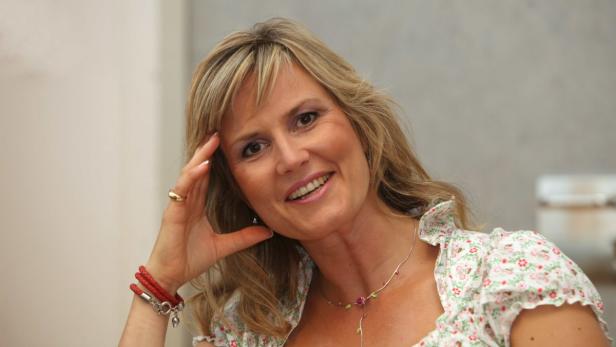 Eine lächelnde Frau mit blonden Haaren und einem geblümten Oberteil posiert für ein Porträt.