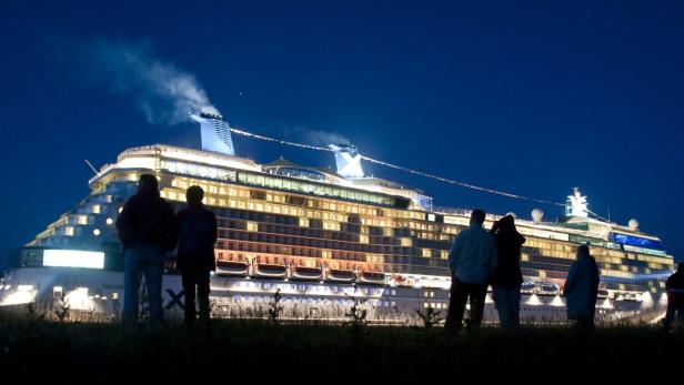 Eine Gruppe von Menschen betrachtet nachts ein großes, hell erleuchtetes Kreuzfahrtschiff.