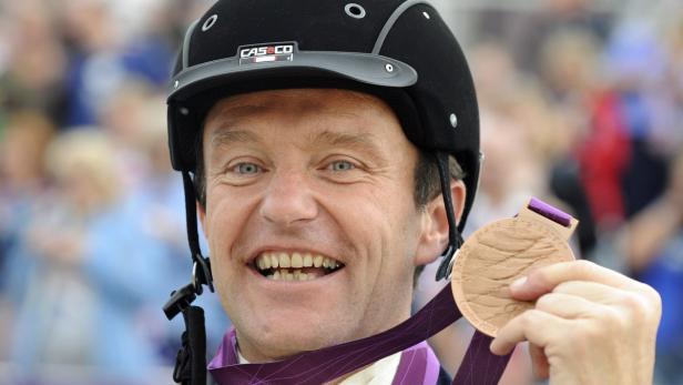 Ein lächelnder Mann mit Reithelm hält eine Bronzemedaille der Olympischen Spiele 2012 in London.