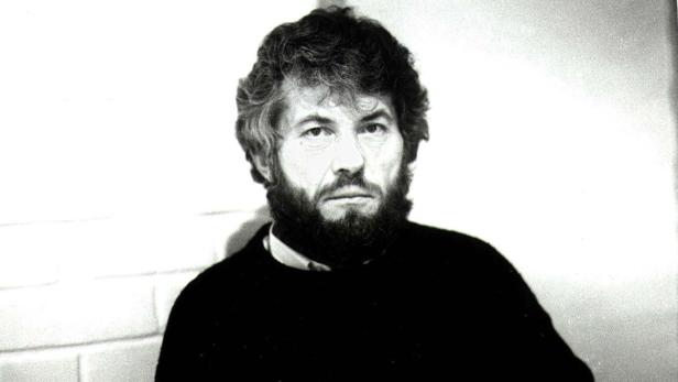 Ein Schwarzweißporträt eines Mannes mit Bart und dunklem Pullover vor einer hellen Wand.