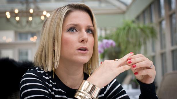 Eine Frau mit blonden Haaren, gestreiftem Oberteil und goldenem Armreif sitzt in einem hellen Raum.