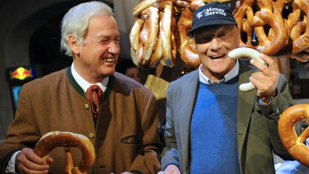 Zwei Männer halten Brezeln und eine Weißwurst vor einem Hintergrund von Brezeln.