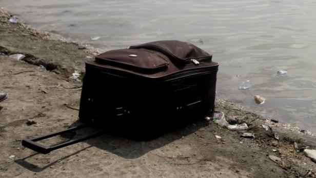 Ein brauner Rollkoffer steht am Ufer eines Gewässers, umgeben von Müll.