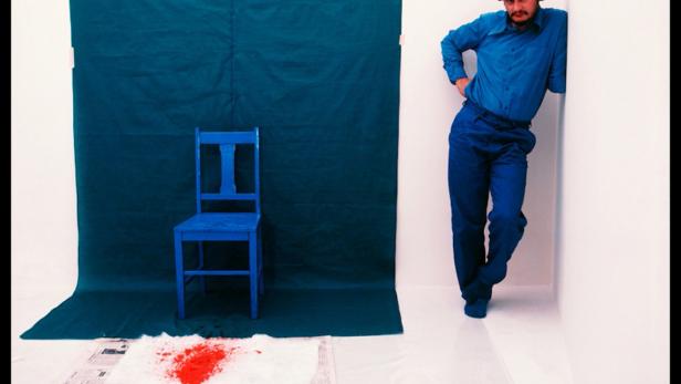 Ein Mann in blauer Kleidung lehnt an einer Wand; im Raum stehen ein blauer Stuhl und eine Zeitung mit roter Farbe.