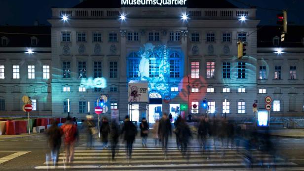 Das MuseumsQuartier in Wien ist nachts beleuchtet, Menschen überqueren einen Zebrastreifen.