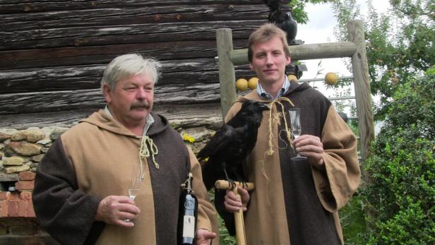 Zwei Männer in Kutten posieren mit einem Raben und Weingläsern vor einem Holzhaus.