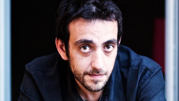 Porträt eines Mannes mit dunklen Haaren und Bart vor einem dunklen Hintergrund.