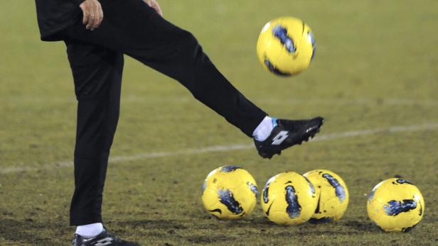 Ein Mann in Anzughose und Fußballschuhen jongliert einen gelben Fußball.