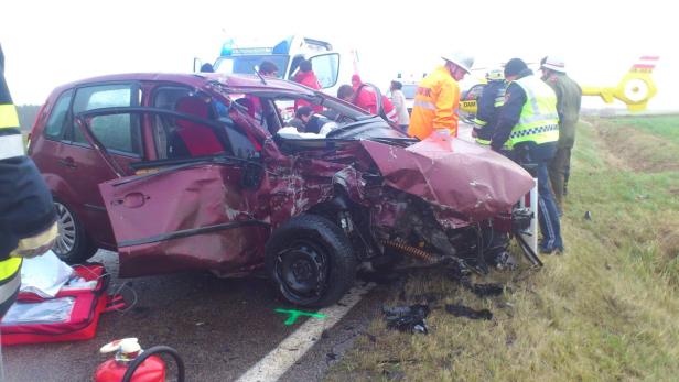 Ein schwer beschädigtes rotes Auto nach einem Unfall mit Rettungskräften und einem Hubschrauber vor Ort.