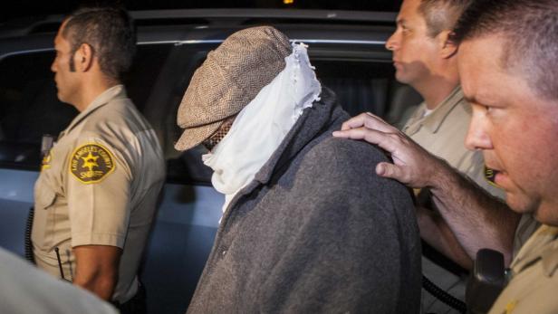 Eine Person mit Hut und Verbänden wird von Polizisten des Los Angeles County begleitet.