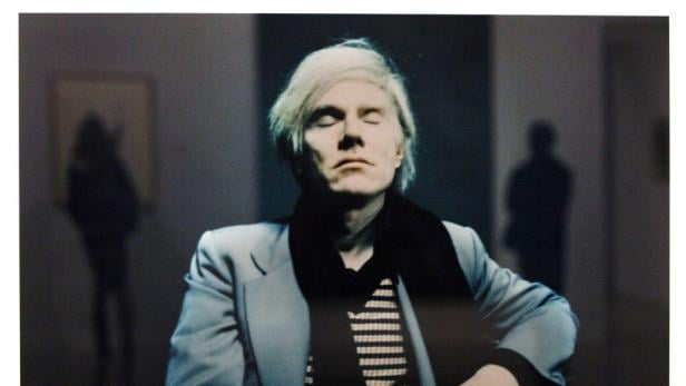 Andy Warhol mit geschlossenen Augen, einen Schal und einen hellen Blazer tragend.