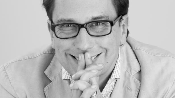 Ein lächelnder Mann mit Brille und hellem Sakko in Schwarzweiß.