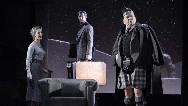 Szene aus einer Theateraufführung mit drei Schauspielern und Gepäck vor einer projizierten Schneelandschaft.