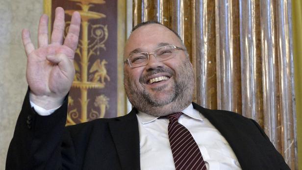 Ein lächelnder Mann mit Brille hebt seine Hand mit vier ausgestreckten Fingern.