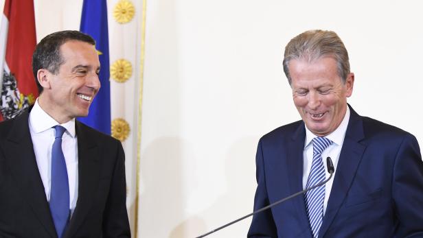 Zwei Männer in Anzügen stehen vor Flaggen und lächeln.