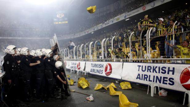 Polizisten mit Schutzschilden stehen vor tobenden Fußballfans in einem Stadion.