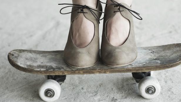 Ein Paar Füße in eleganten Schuhen stehen auf einem abgenutzten Skateboard.