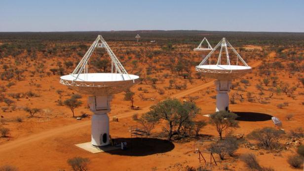 Mehrere Radioteleskope stehen in einer trockenen, baumbestandenen Landschaft.