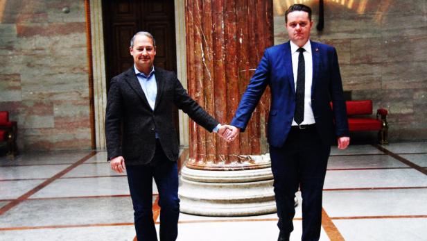 Zwei Männer in Anzügen geben sich vor einer Säule die Hand.