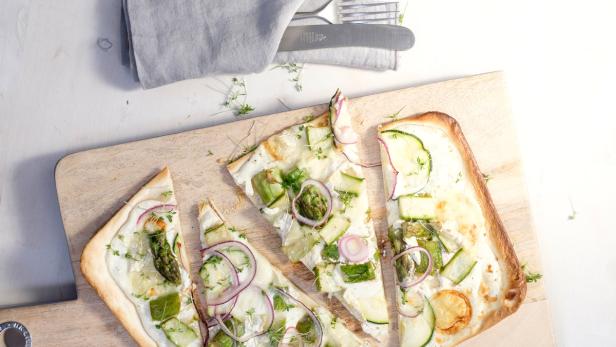 Ein Flammkuchen mit Gemüse wie Spargel, Zucchini und roten Zwiebeln auf einem Holzbrett.