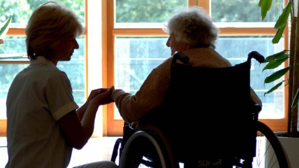 Eine ältere Frau im Rollstuhl hält die Hand einer jüngeren Frau.