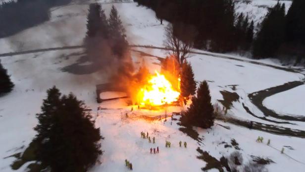 Ein Haus steht in einer verschneiten Landschaft in Flammen, während Feuerwehrleute zusehen.