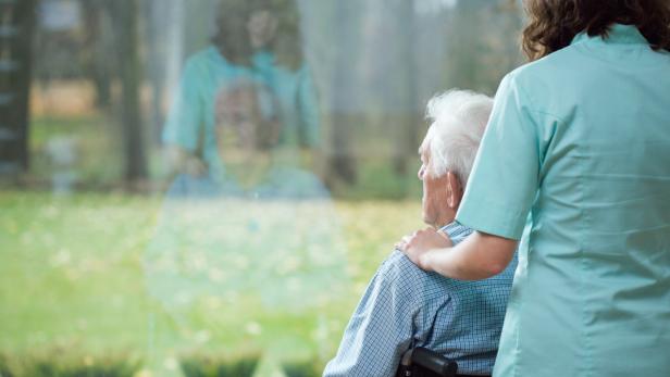 Eine Krankenschwester legt ihre Hand auf die Schulter eines älteren Mannes im Rollstuhl.