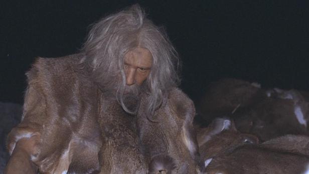 Ein Mann mit langem, grauem Haar und Fellkleidung blickt nach unten.