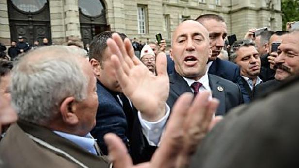 Ein Mann in Anzug gestikuliert inmitten einer Menschenmenge vor einem Gebäude.