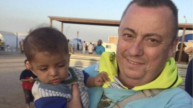 Ein Mann hält ein Baby im Arm, im Hintergrund Zelte und andere Menschen.