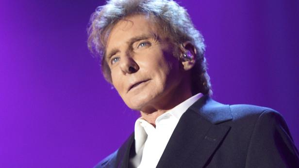 Barry Manilow in einem schwarzen Anzug vor einem violetten Hintergrund.