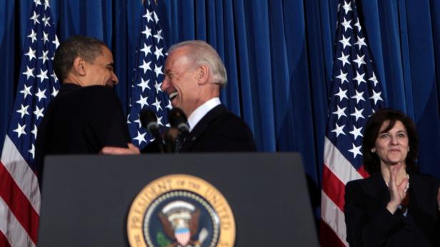 Barack Obama und Joe Biden stehen lachend vor amerikanischen Flaggen.