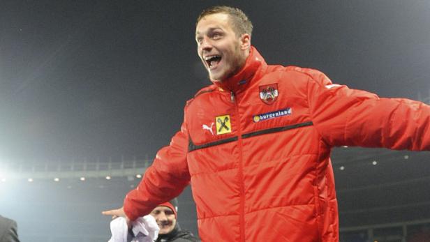 Ein jubelnder österreichischer Fußballspieler in roter Jacke mit ausgebreiteten Armen.