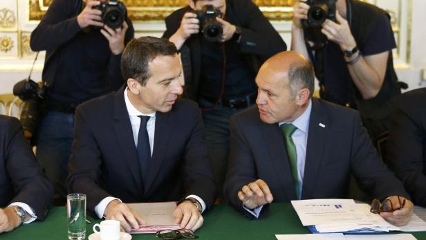 Zwei Männer in Anzügen unterhalten sich an einem Tisch, umringt von Fotografen.