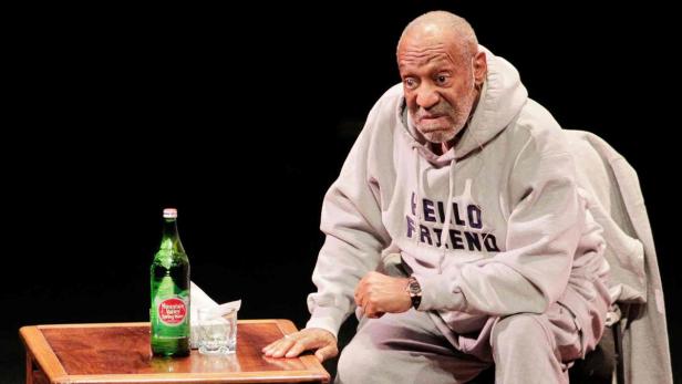 Bill Cosby sitzt auf der Bühne neben einem Tisch mit Wasserflasche und Glas.
