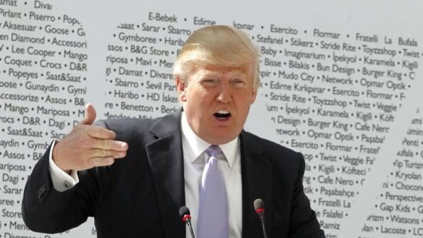 Donald Trump gestikuliert vor einer Wand mit Markennamen.
