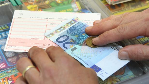 Eine Hand bezahlt mit einem 20-Euro-Schein und einer Münze einen Lottoschein.