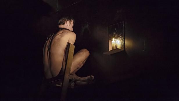 Ein Mann sitzt gefesselt auf einem Stuhl, im Hintergrund Kerzenlicht.