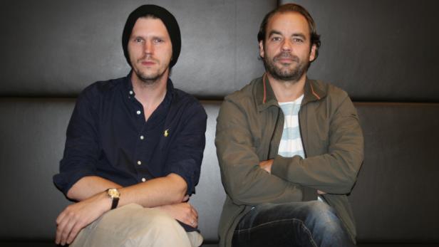 Zwei Männer sitzen mit verschränkten Armen auf einer Bank vor einer dunklen Wand.