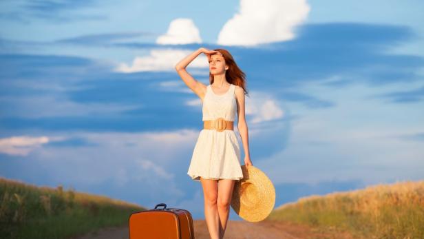 Eine junge Frau mit Koffer und Hut steht auf einem Feldweg und schaut in die Ferne.