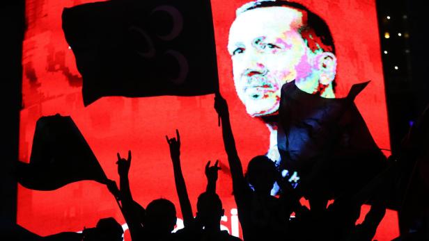 Eine Menschenmenge jubelt vor einem roten Bildschirm mit dem Porträt von Recep Tayyip Erdoğan.