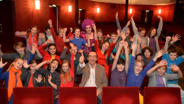 Eine Gruppe Kinder und Erwachsene jubeln in einem Theater, darunter eine Dragqueen mit lila Perücke.