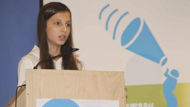 Ein junges Mädchen hält eine Rede an einem Rednerpult vor einem blauen Logo.