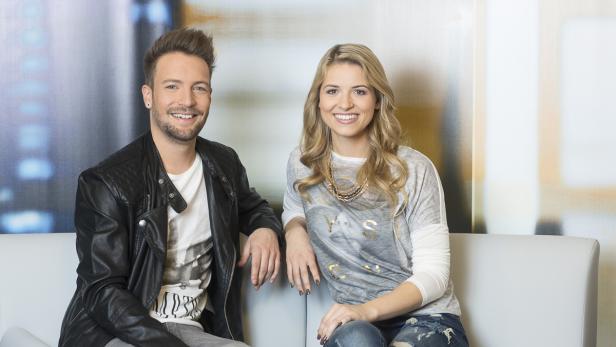 Ein Mann mit Lederjacke und eine Frau mit Halskette sitzen lächelnd auf einem Sofa.