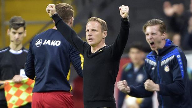 Ein Fußballtrainer jubelt mit erhobenen Fäusten am Spielfeldrand.