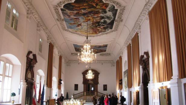 Ein großer Saal mit Deckenmalereien und Kristalllüstern, in dem sich Menschen aufhalten.